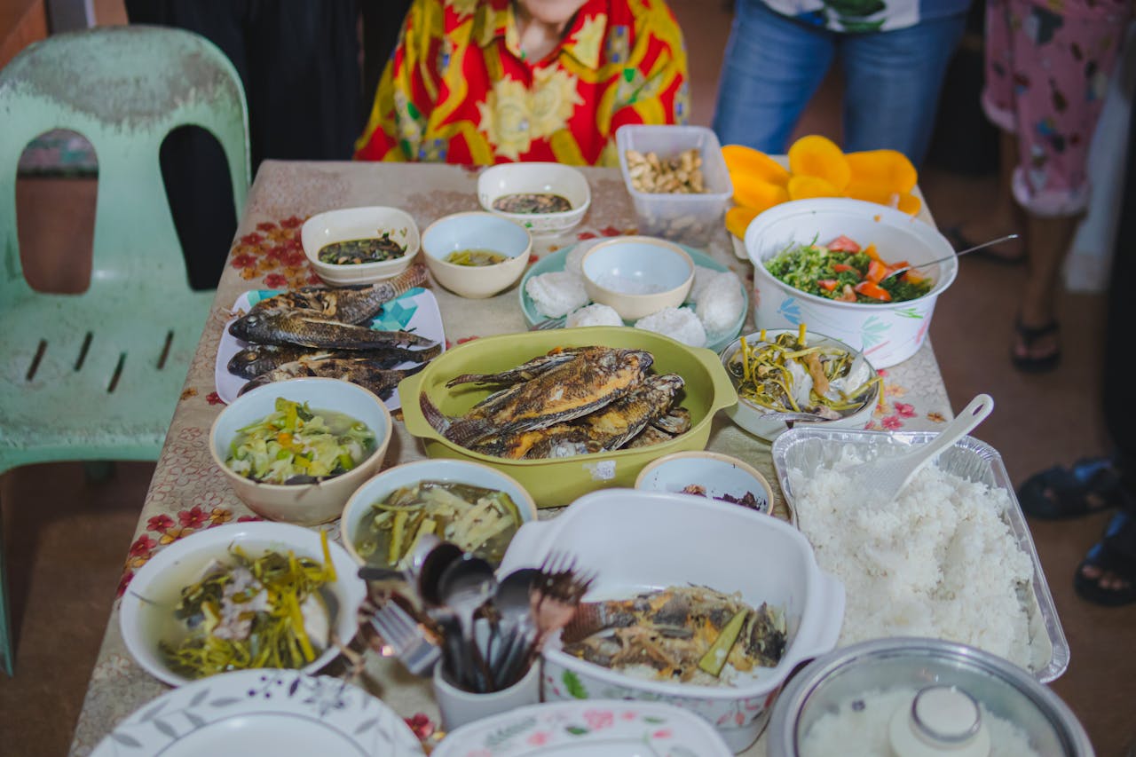 A Filipino family gathering with a table full of traditional home-cooked dishes, including grilled fish, rice, vegetables, and soup.
