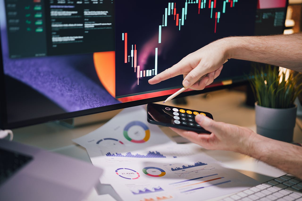 A person analyzing stock market trends on a computer screen with candlestick charts and data while using a calculator and financial reports.